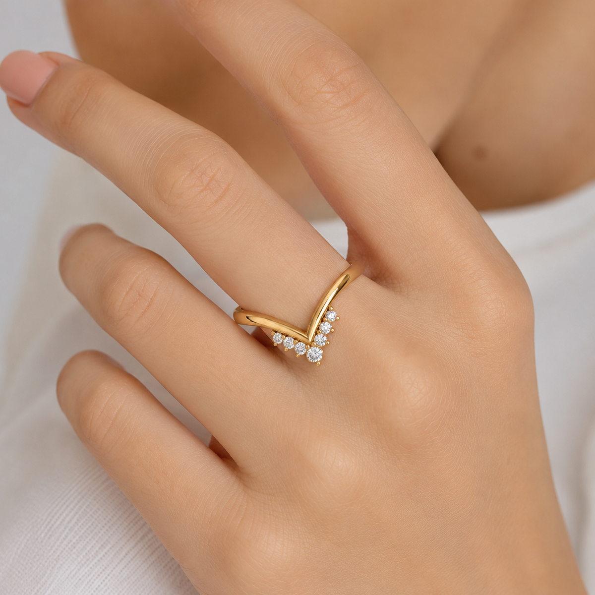 A close-up of a hand wearing a gold V-shaped ring with small clear stones along the front, set against a soft white background.