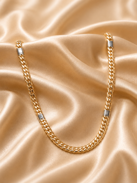 A gold-toned Cuban link chain necklace with silver-toned rectangular end caps, draped on a shiny champagne satin fabric background.