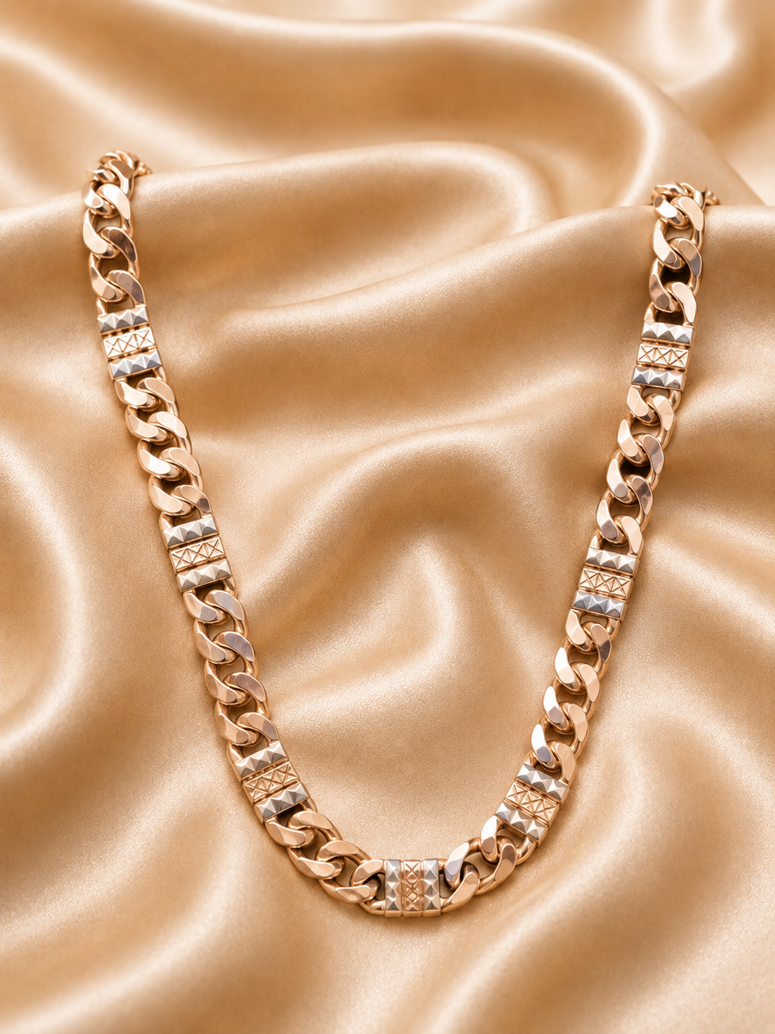 A chunky gold-toned Cuban link chain necklace with rectangular links, displayed on a draped champagne satin fabric background.