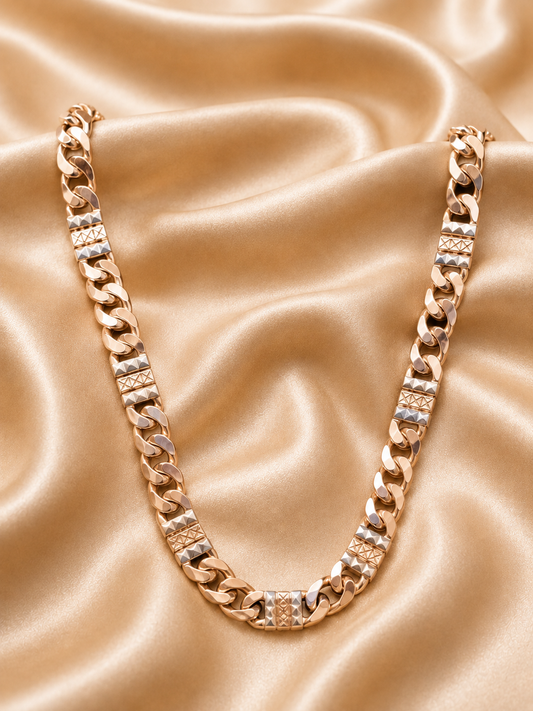 A chunky gold-toned Cuban link chain necklace with rectangular links, displayed on a draped champagne satin fabric background.