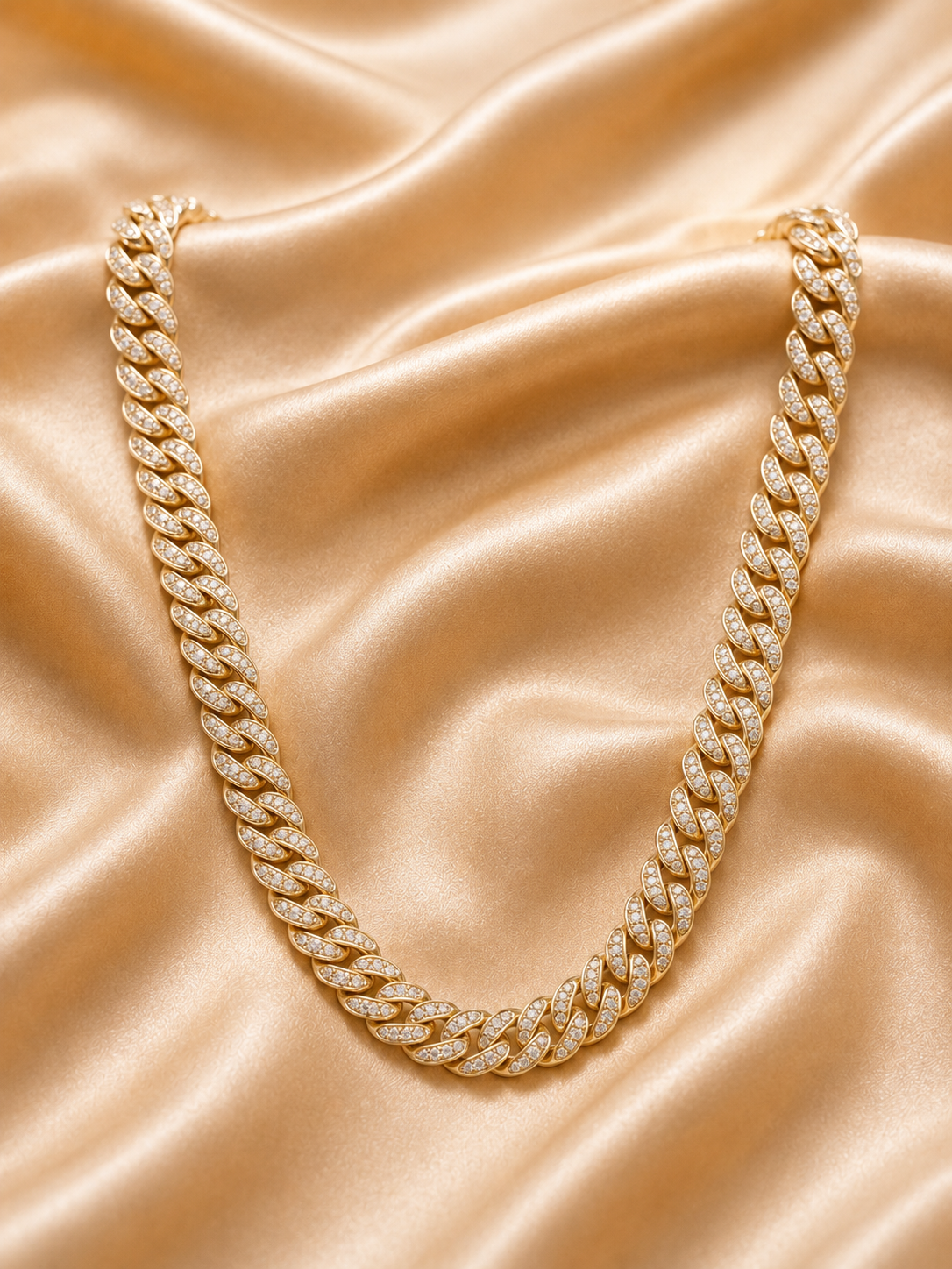 A gold Cuban link chain necklace paved with small clear stones, arranged in a U-shape on a draped champagne satin fabric background.