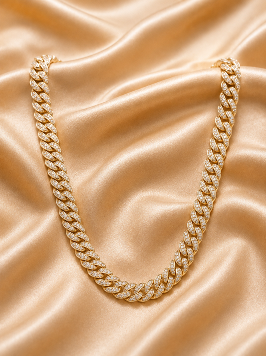 A gold Cuban link chain necklace paved with small clear stones, arranged in a U-shape on a draped champagne satin fabric background.