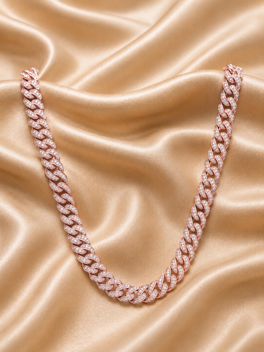 A rose gold Cuban link chain necklace paved with small clear stones, arranged in a U-shape on a draped champagne satin fabric background.