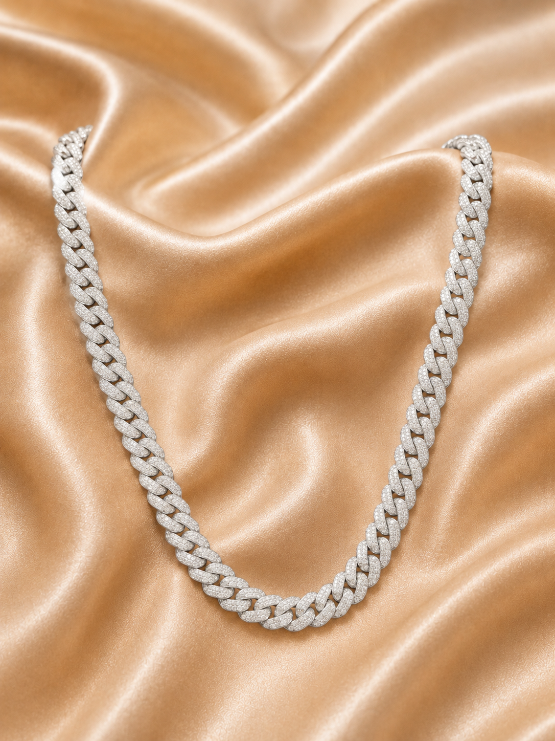 A silver-toned Cuban link chain necklace, heavily embellished with clear stones, arranged in a U-shape on a draped champagne-colored satin fabric.