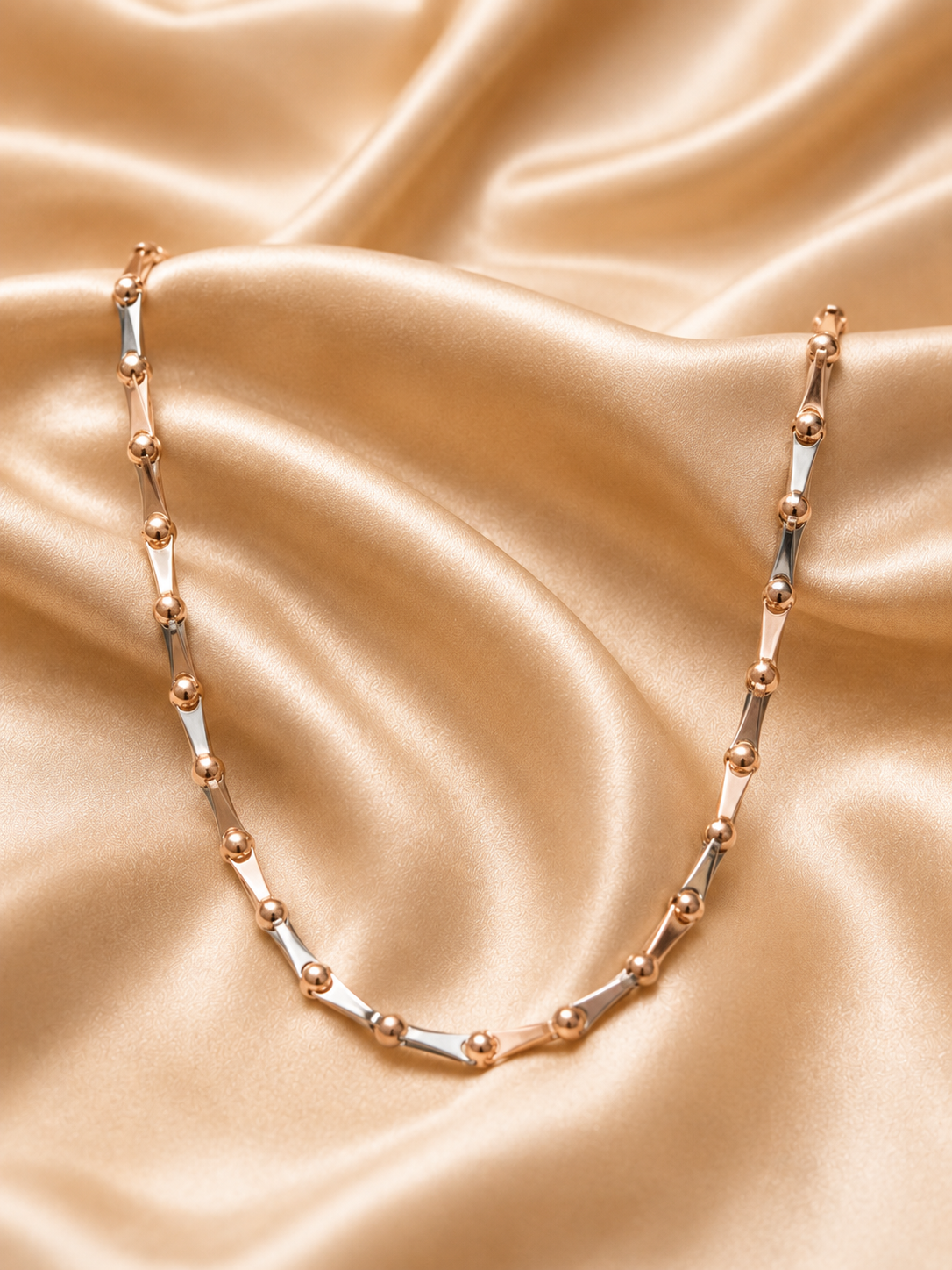 A delicate chain necklace with alternating polished rose gold and silver cylindrical links and small round beads, displayed on draped champagne satin fabric.