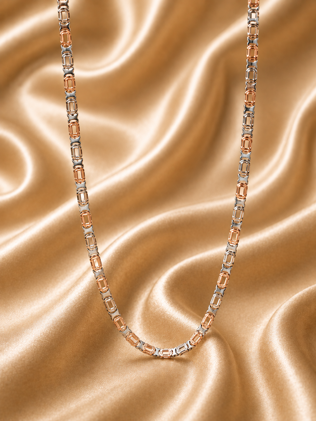 A two-tone chain necklace featuring rose gold and silver-toned metal links, accented with rectangular clear gemstones, displayed on a draped champagne satin fabric background.