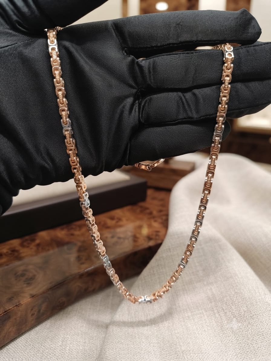 A close-up of a delicate chain necklace with alternating rose gold and silver links, held by a hand wearing a black satin glove over a light beige fabric surface.