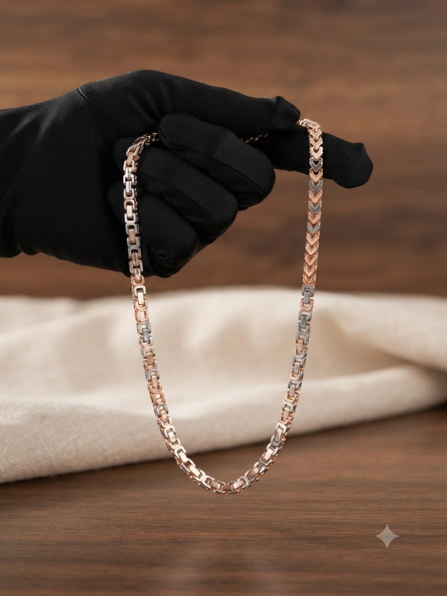 A close-up of a black-gloved hand holding a rose gold chain necklace with alternating rose gold and silver-toned links, set against a warm wooden background with a soft beige cloth.